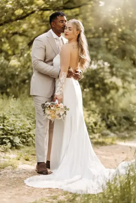 Eine Braut in einem weißen Kleid und ein Bräutigam in einem hellbeigen Anzug umarmen sich im Freien auf einem sonnenbeschienenen, begrünten Weg - wunderschön eingefangen von Hochzeitsfotograf Görlitz auf Hochzeit Gut am See.