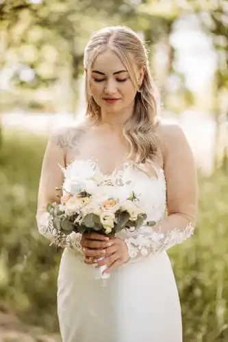 Eine Braut mit langen blonden Haaren in einem weißen Spitzenhochzeitskleid hält einen Strauß blasser Blumen. Sie steht im Freien auf der Hochzeit Gut am See, die Sonne scheint und das Grün leuchtet, aufgenommen von Hochzeitsfotograf Görlitz.