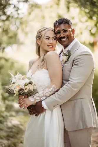 Eine Braut in weißem Kleid und Blumenstrauß steht lächelnd mit ihrem Bräutigam in einem hellbeigen Anzug und Brille, beide umarmen sich in einer sonnenbeschienenen, grünen Umgebung - perfekte Momente für jeden Hochzeitsfotografen Görlitz auf einer Hochzeit Gut am See.