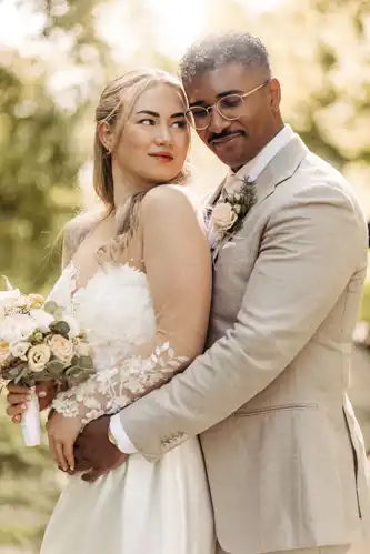 Eine Braut in einem weißen Spitzenhochzeitskleid hält einen Blumenstrauß, während sie von einem Bräutigam in einem beigen Anzug umarmt wird; beide lächeln sanft in den sonnenbeschienenen Gärten von Hochzeit Gut am See, aufgenommen von Hochzeitsfotograf Görlitz.