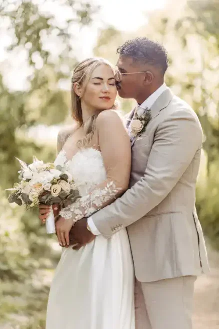 Eine Braut in einem weißen Kleid hält einen Blumenstrauß, während sie mit ihrem Bräutigam in einem hellbeigen Anzug steht. Sie umarmen sich im Freien auf einem sonnenbeschienenen, grünen Weg auf Hochzeit Gut am See, umgeben von Bäumen und Laub. Hochzeitsfotograf Görlitz hat den Moment festgehalten.