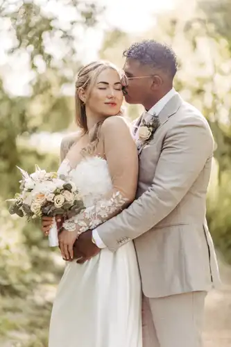 Eine Braut in einem weißen Kleid hält einen Blumenstrauß, während sie mit ihrem Bräutigam in einem hellbeigen Anzug steht. Sie umarmen sich im Freien auf einem sonnenbeschienenen, grünen Weg auf Hochzeit Gut am See, umgeben von Bäumen und Laub. Hochzeitsfotograf Görlitz hat den Moment festgehalten.