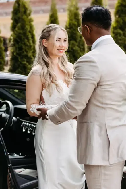 Eine Braut in einem weißen Kleid lächelt einen Bräutigam in einem hellen Anzug an, während sie neben einem schwarzen Auto im Freien auf Hochzeit Gut am See stehen, mit grünen Bäumen im Hintergrund. Aufgenommen von Hochzeitsfotograf Görlitz.