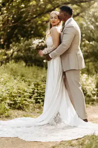 Eine Braut in einem weißen Kleid hält einen Blumenstrauß, während sie mit ihrem Bräutigam in einem hellbeigen Anzug steht. Sie umarmen sich im Freien auf einem sonnenbeschienenen, grünen Weg auf Hochzeit Gut am See, umgeben von Bäumen und Laub. Hochzeitsfotograf Görlitz hat den Moment festgehalten.