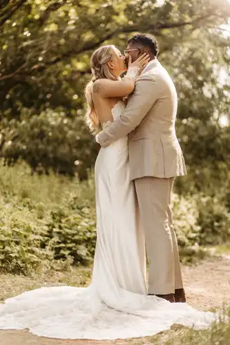 Eine Braut in einem weißen Kleid und ein Bräutigam in einem hellen Anzug umarmen und küssen sich im Freien auf einem sonnenbeschienenen, begrünten Weg auf Hochzeit Gut am See, umgeben von Grün. Wunderschön eingefangen von Hochzeitsfotograf Görlitz.