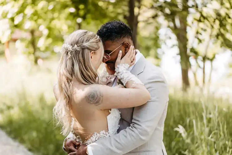 Eine Braut und ein Bräutigam umarmen sich eng im Freien auf der Hochzeit Gut am See, umgeben von Grün. Die Braut trägt ein weißes Spitzenkleid mit offenem Rücken, der Bräutigam einen hellen Anzug und eine Sonnenbrille. Sie lächeln und berühren sich liebevoll an der Stirn.