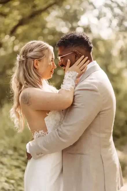 Braut und Bräutigam umarmen sich im Freien auf der Hochzeit Gut am See und blicken sich liebevoll in die Augen. Die Braut trägt ein weißes Kleid mit Spitzenärmeln, der Bräutigam einen hellbraunen Anzug. Grünzeug und Sonnenlicht füllen den Hintergrund. Hochzeitsfotograf Görlitz.