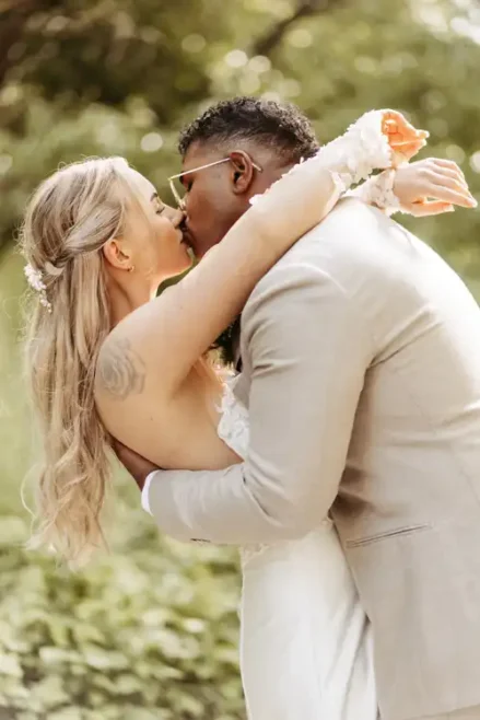 Eine Braut und ein Bräutigam geben sich einen romantischen Kuss im Freien auf der Hochzeit Gut am See, umgeben von viel Grün. Die Braut trägt ein weißes Kleid mit Blumen im Haar, während der Bräutigam in einem hellen Anzug und mit Brille diesen magischen Moment vervollständigt, der von Hochzeitsfotograf Görlitz festgehalten wurde.