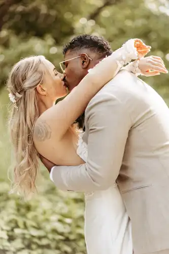 Eine Braut und ein Bräutigam geben sich einen romantischen Kuss im Freien auf der Hochzeit Gut am See, umgeben von viel Grün. Die Braut trägt ein weißes Kleid mit Blumen im Haar, während der Bräutigam in einem hellen Anzug und mit Brille diesen magischen Moment vervollständigt, der von Hochzeitsfotograf Görlitz festgehalten wurde.