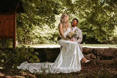 Eine Braut in einem weißen Kleid sitzt auf einer Steinmauer, hält einen Blumenstrauß in der Hand und lächelt den Bräutigam im hellen Anzug an, umgeben von Grün und Bäumen auf Hochzeit Gut am See. Ein unvergesslicher Moment unter freiem Himmel, festgehalten von Hochzeitsfotograf Görlitz.