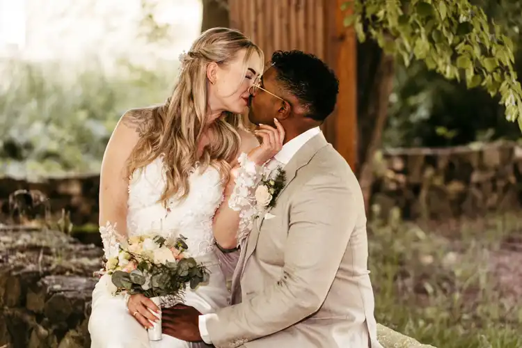 Eine Braut in einem weißen Kleid und ein Bräutigam in einem beigen Anzug sitzen eng beieinander auf einem Steinsims in Hochzeit Gut am See, berühren sich sanft und sind im Begriff, sich zu küssen. Die Braut hält einen Blumenstrauß in der Hand, beide wirken glücklich und verliebt. Hochzeitsfotograf Görlitz hat den Moment festgehalten.
