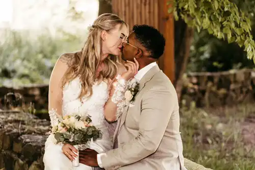 Eine Braut in einem weißen Kleid und ein Bräutigam in einem beigen Anzug sitzen eng beieinander auf einem Steinsims in Hochzeit Gut am See, berühren sich sanft und sind im Begriff, sich zu küssen. Die Braut hält einen Blumenstrauß in der Hand, beide wirken glücklich und verliebt. Hochzeitsfotograf Görlitz hat den Moment festgehalten.