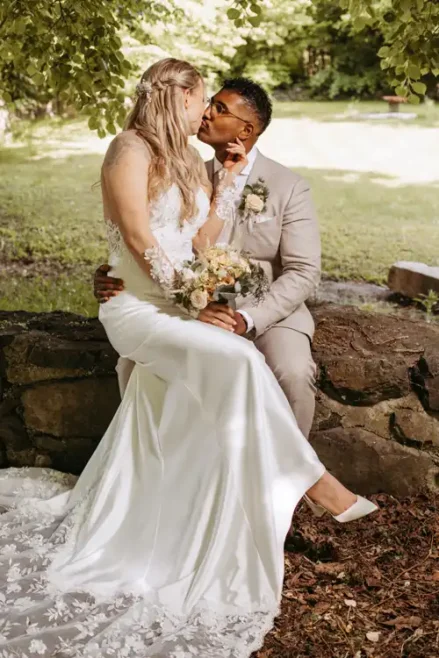 Eine Braut in einem weißen Kleid und ein Bräutigam in einem hellen Anzug sitzen auf einer Steinmauer bei Hochzeit Gut am See, halten einen Blumenstrauß und schauen sich unter grünen Bäumen in die Augen - aufgenommen von Hochzeitsfotograf Görlitz in der sonnigen Natur.