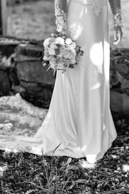 Eine Braut in einem langen, eleganten Kleid hält einen Blumenstrauß an ihrer Seite. Das von Hochzeitsfotograf Görlitz aufgenommene Schwarz-Weiß-Foto zeigt nur ihren Unterkörper und ihr Kleid, während eine Steinmauer und verstreute Blätter auf die Hochzeit Gut am See hindeuten.
