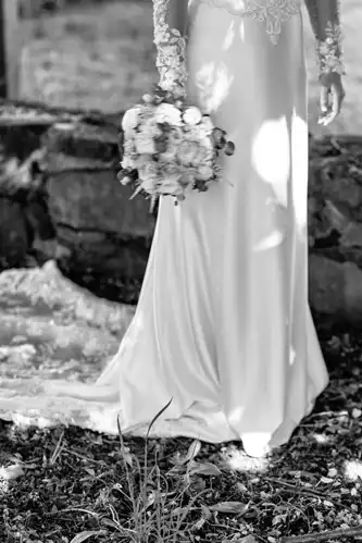 Eine Braut in einem langen, eleganten Kleid hält einen Blumenstrauß an ihrer Seite. Das von Hochzeitsfotograf Görlitz aufgenommene Schwarz-Weiß-Foto zeigt nur ihren Unterkörper und ihr Kleid, während eine Steinmauer und verstreute Blätter auf die Hochzeit Gut am See hindeuten.