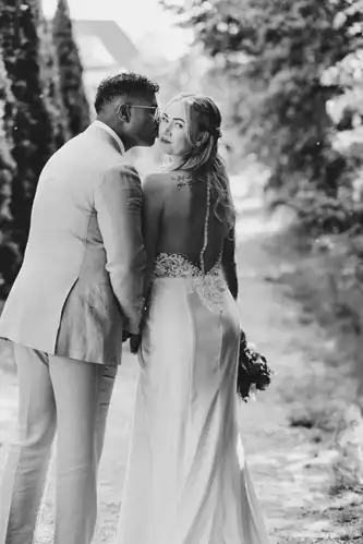 Ein Bräutigam in einem hellen Anzug küsst die Wange der Braut, die in einem rückenfreien Kleid und mit einem Blumenstrauß in der Hand auf einem von Bäumen gesäumten Weg auf Hochzeit Gut am See steht, während sie in die Kamera schaut.