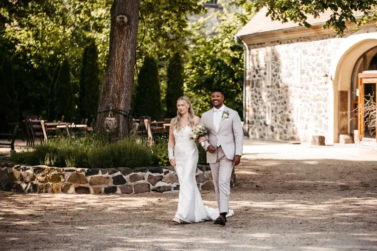 Eine Braut in einem weißen Kleid und ein Bräutigam in einem hellen Anzug halten sich an den Händen und lächeln, während sie an einem sonnigen Tag im Freien auf Hochzeit Gut am See spazieren gehen, mit Bäumen und einem Steingebäude im Hintergrund.