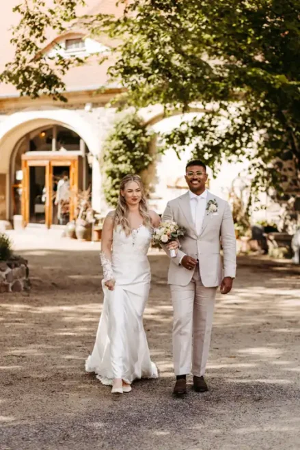 Eine Braut in einem weißen Kleid und ein Bräutigam in einem hellen Anzug spazieren Arm in Arm im Freien, lächelnd, mit Bäumen und dem charmanten Hochzeitsgut am See im Hintergrund.