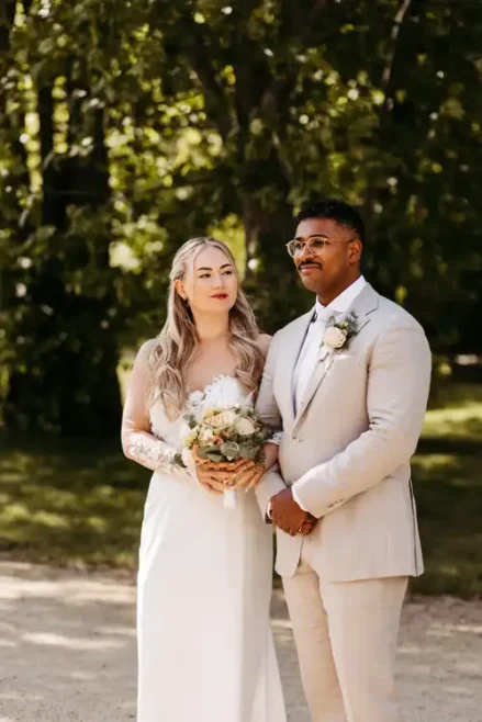 Eine Braut in einem weißen Kleid mit einem Blumenstrauß in der Hand steht neben einem Bräutigam in einem hellen Anzug und einer Brille. Draußen auf Hochzeit Gut am See posieren sie im Grünen für ein Hochzeitsfoto von Hochzeitsfotograf Görlitz.