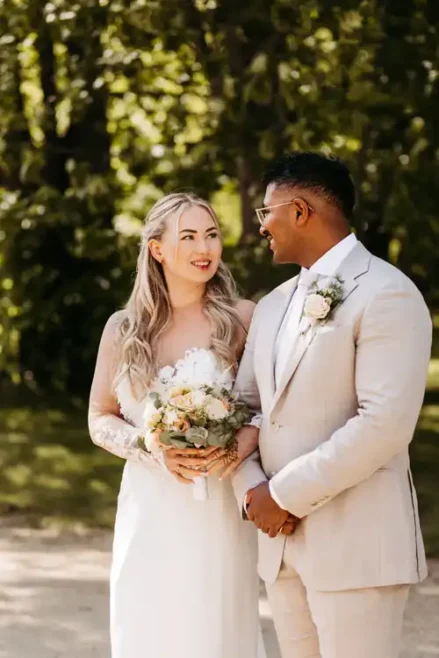Eine Braut in einem weißen Kleid, die einen Blumenstrauß hält, steht neben einem Bräutigam in einem hellen Anzug. Sie lächeln sich im Grünen auf Hochzeit Gut am See an, fotografiert von Hochzeitsfotograf Görlitz.