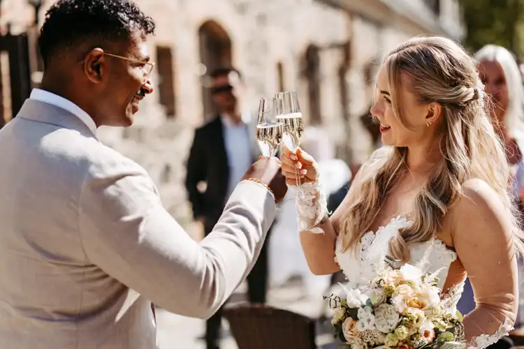 Eine Braut und ein Bräutigam lächeln und stoßen im Freien auf ihrer Hochzeit auf Gut am See mit Sekt an. Die Braut hält einen Blumenstrauß in der Hand, und beide sind formell gekleidet, während im Hintergrund Gäste zu sehen sind.