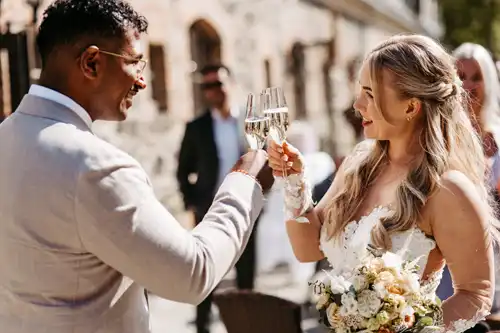 Eine Braut und ein Bräutigam lächeln und stoßen im Freien auf ihrer Hochzeit auf Gut am See mit Sekt an. Die Braut hält einen Blumenstrauß in der Hand, und beide sind formell gekleidet, während im Hintergrund Gäste zu sehen sind.