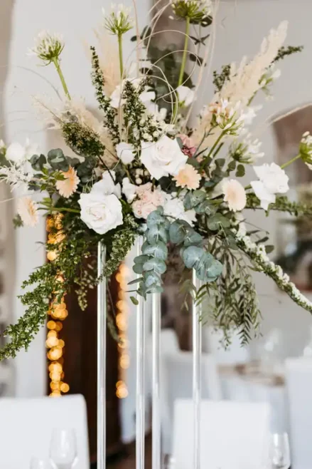 Großes Blumenarrangement in Glasvasen mit weißen Rosen, blassrosa Blumen, Grünzeug und langen Ziergräsern - perfekt für eine Hochzeit auf Gut am See, vor einem sanft beleuchteten Innenhintergrund mit verschwommenen Lichtern.