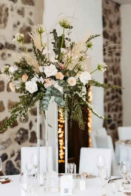 Hoher Blumenschmuck mit weißen Rosen, Grünzeug und beigen Blumen auf einem durchsichtigen Ständer bei Hochzeit Gut am See; eleganter Tisch mit Glaswaren und weißen Stühlen unter warmen Lichterketten - wunderschön eingefangen von Hochzeitsfotograf Görlitz.