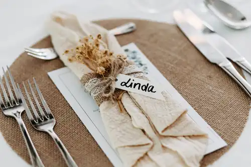 Eine rustikale Tischdekoration auf der Hochzeit Gut am See besteht aus einer gefalteten cremefarbenen Serviette, die mit Schnur, getrockneten Blumen, Spitzenband und einem "Linda"-Namensschild auf einem Platzdeckchen aus Sackleinen gebunden ist, wunderschön festgehalten von Hochzeitsfotograf Görlitz.