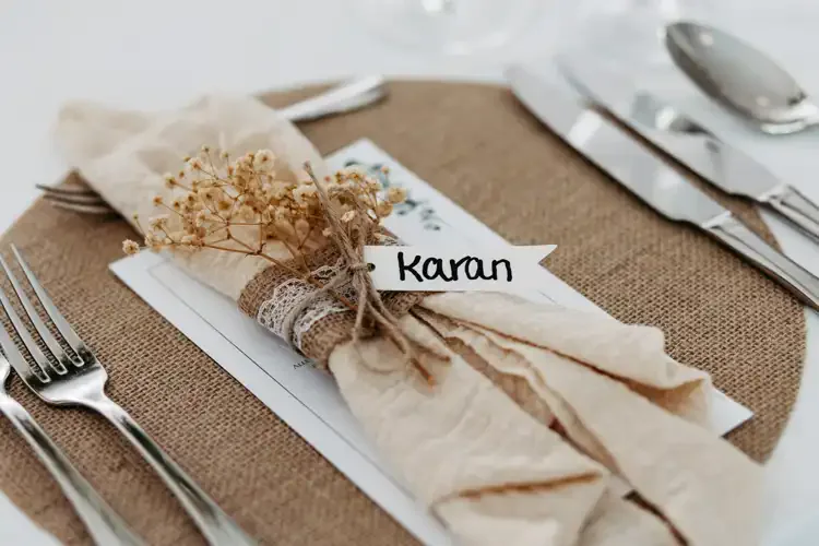 Auf dem rustikalen Tisch der Hochzeit Gut am See liegt eine beigefarbene Serviette mit Schnur und Spitze umwickelt und mit Trockenblumen geschmückt auf einem Platzset aus Jute. Ein Namenskärtchen mit der Aufschrift Karan ist angebracht, umgeben von Silberbesteck.