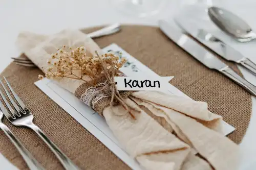 Auf dem rustikalen Tisch der Hochzeit Gut am See liegt eine beigefarbene Serviette mit Schnur und Spitze umwickelt und mit Trockenblumen geschmückt auf einem Platzset aus Jute. Ein Namenskärtchen mit der Aufschrift Karan ist angebracht, umgeben von Silberbesteck.