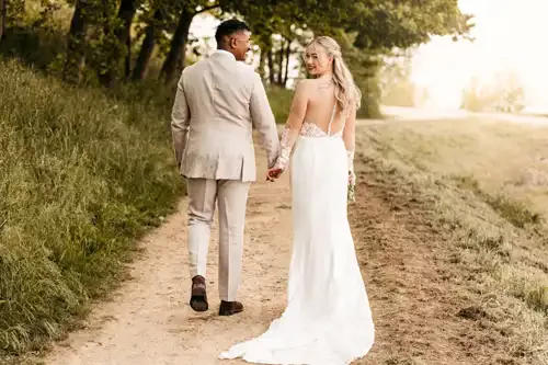 Eine Braut und ein Bräutigam gehen Hand in Hand einen von Grün umgebenen Feldweg auf Hochzeit Gut am See hinunter. Die Braut trägt ein weißes Kleid mit langer Schleppe und Ärmeln und lächelt zurück, während der Bräutigam in einem beigen Anzug neben ihr hergeht. Hochzeitsfotograf Görlitz hält den Moment fest.