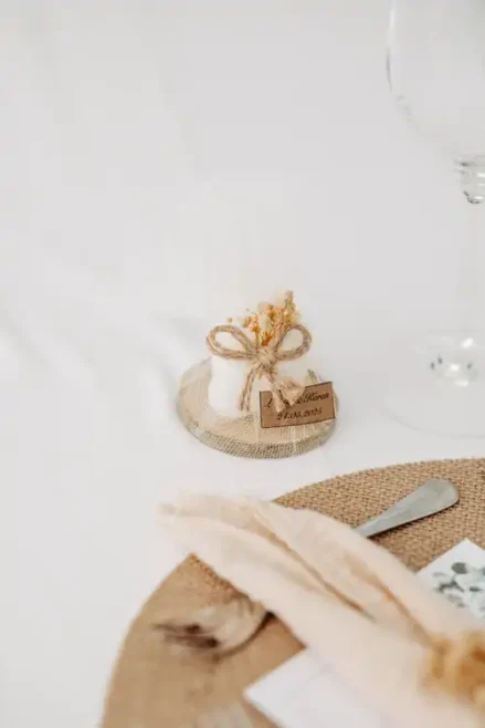Ein kleines, in weißes Tuch eingewickeltes und mit Schnur und getrockneten Blumen verschnürtes Hochzeitsgeschenk liegt auf einem Untersetzer aus Sackleinen, wunderschön eingefangen von Hochzeitsfotograf Görlitz, neben einem Gedeck mit beiger Serviette und Geschirr auf einem gewebten Tischset.