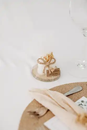 Ein kleines, in weißes Tuch eingewickeltes und mit Schnur und getrockneten Blumen verschnürtes Hochzeitsgeschenk liegt auf einem Untersetzer aus Sackleinen, wunderschön eingefangen von Hochzeitsfotograf Görlitz, neben einem Gedeck mit beiger Serviette und Geschirr auf einem gewebten Tischset.