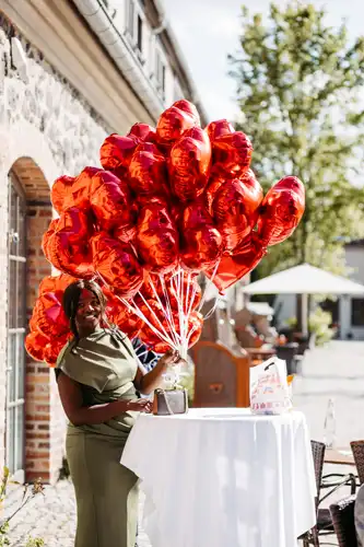 Eine Frau in einem grünen Kleid steht im Freien an einem runden Tisch, hält rote herzförmige Luftballons und lächelt. Der sonnige Hof mit seinen steinernen Gebäuden und den Grünanlagen ist die perfekte Kulisse für eine Hochzeit auf Gut am See.