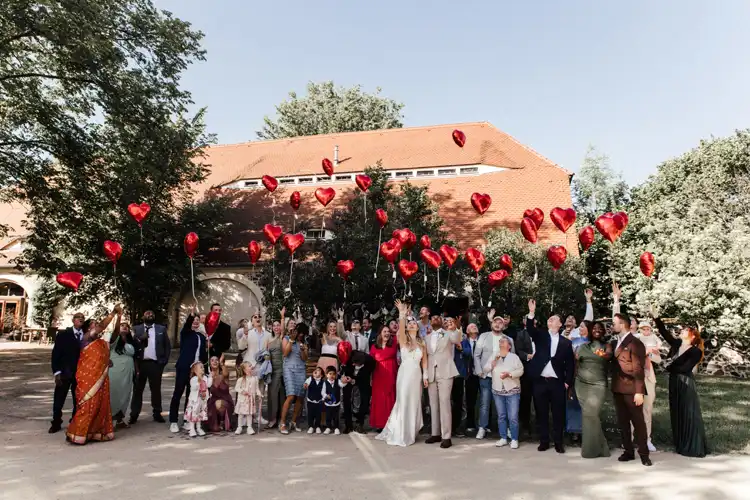 Eine große Hochzeitsgesellschaft steht im Freien auf Hochzeit Gut am See, lächelt und lässt rote herzförmige Luftballons steigen. Die verschiedenen Gäste in formeller und farbenfroher Kleidung versammeln sich freudig vor einem Gebäude mit einem roten Dach. Hochzeitsfotograf Görlitz hält den Moment fest.