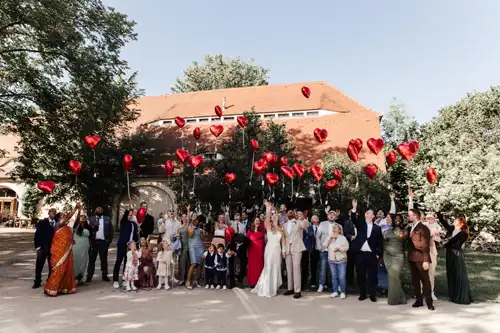 Eine große Hochzeitsgesellschaft steht im Freien auf Hochzeit Gut am See, lächelt und lässt rote herzförmige Luftballons steigen. Die verschiedenen Gäste in formeller und farbenfroher Kleidung versammeln sich freudig vor einem Gebäude mit einem roten Dach. Hochzeitsfotograf Görlitz hält den Moment fest.
