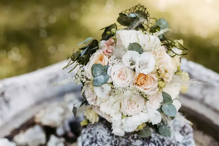 Ein Strauß aus cremefarbenen und zartrosa Rosen gemischt mit weißen Blumen, Eukalyptusblättern und Grünzeug wird auf einer Steinfläche im Freien arrangiert - perfekt für Ihre Hochzeit auf Gut am See und wunderschön festgehalten von Hochzeitsfotograf Görlitz.