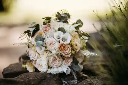 Ein Strauß aus pastellfarbenen Rosen, weißen Blumen und grünem Laub ruht auf Steinen im Freien, sanft beleuchtet von natürlichem Sonnenlicht - perfekt für eine Hochzeit auf Gut am See und wunderschön eingefangen von einem Hochzeitsfotograf Görlitz.