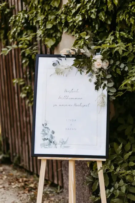 Ein Hochzeitsbegrüßungsschild auf einer Staffelei, geschmückt mit Grünzeug und einer Blume, steht im Freien neben einem Holzzaun und grünen Pflanzen. Auf dem Schild steht auf Deutsch: Herzlich willkommen zu unserer Hochzeit Linda & Karan - Hochzeit Gut am See.