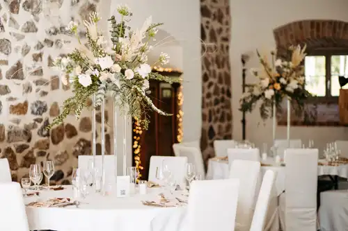 Elegantes Hochzeitszimmer in der Hochzeit Gut am See mit runden Tischen mit weißen Tüchern, weißen Stühlen und hohen Blumensträußen. Steinwände und sanftes Licht schaffen eine warme Atmosphäre - perfekt, um Erinnerungen mit Hochzeitsfotograf Görlitz festzuhalten.