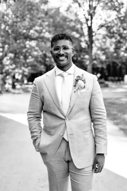 Ein lächelnder Mann mit Brille, hellem Anzug und Krawatte steht im Freien auf der Hochzeit Gut am See, eine Hand in der Tasche. Er hat eine Boutonnière an sein Jackett geheftet, die der Hochzeitsfotograf Görlitz vor einem unscharfen Hintergrund aus Bäumen aufgenommen hat.