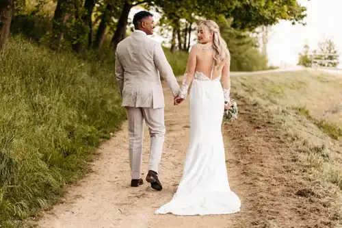 Eine Braut in einem weißen Kleid und ein Bräutigam in einem hellen Anzug gehen Hand in Hand einen von Bäumen gesäumten Feldweg bei Hochzeit Gut am See hinunter. Die Braut blickt über ihre Schulter zurück und lächelt, während sie einen kleinen Blumenstrauß hält, aufgenommen von Hochzeitsfotograf Görlitz.