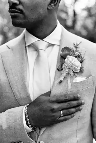 Ein Mann in hellem Anzug und Krawatte, der einen Ring und ein Armband trägt, legt seine Hand auf seine Brust. Eine Rosenboutonniere ziert sein Revers. Aufgenommen in schwarz-weiß von Hochzeitsfotograf Görlitz auf einer Hochzeit im Gut am See.