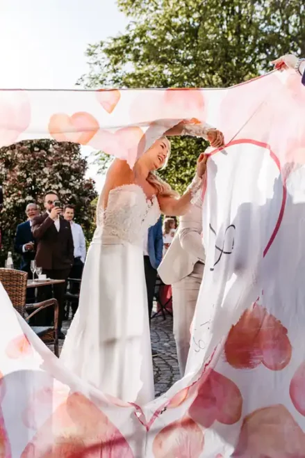 Eine Braut in einem weißen Kleid lächelt, als sie und eine andere Person bei ihrer Hochzeit auf Gut am See ein Herz in ein Laken schneiden. Die Gäste schauen bei dieser fröhlichen Hochzeitsfeier im Freien zu. Hochzeitsfotograf Görlitz hat den Moment wunderschön eingefangen.