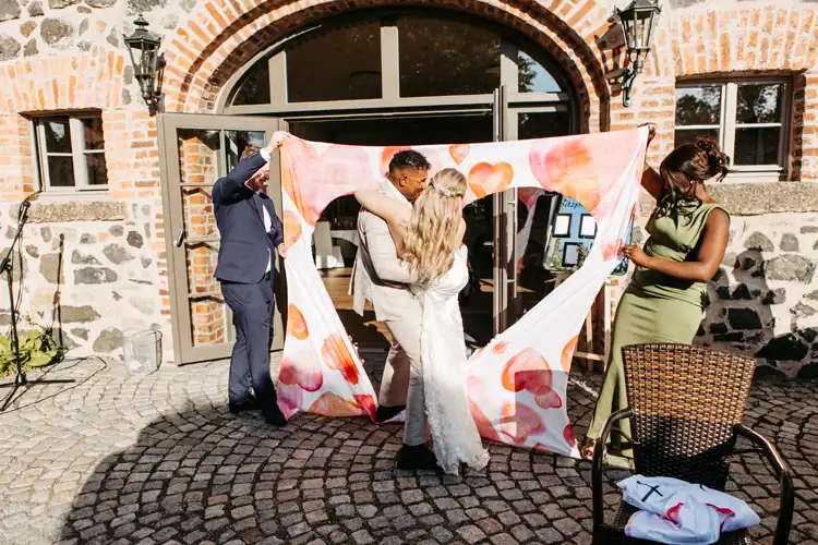 Eine Braut und ein Bräutigam schreiten durch einen herzförmigen Ausschnitt in einem dekorierten Bettlaken, während einer Hochzeit im Freien auf Gut am See auf einer kopfsteingepflasterten Terrasse vor einem Backsteinhaus, wunderschön eingefangen von Hochzeitsfotograf Görlitz.