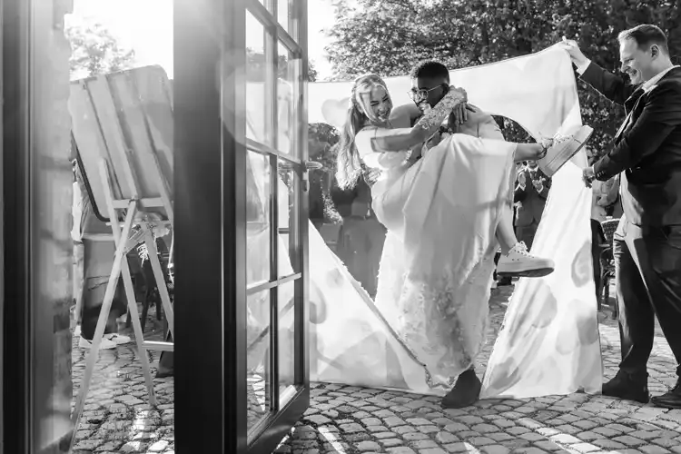 Eine Braut trägt den Bräutigam freudig durch ein ausgeschnittenes herzförmiges Tuch bei ihrer Hochzeit auf Gut am See, während das Sonnenlicht bei dieser Feier im Freien auf das Kopfsteinpflaster fällt.