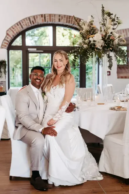 Eine Braut in einem weißen Kleid sitzt auf dem Schoß ihres Bräutigams in einem hellen Anzug bei ihrer Hochzeit auf Gut am See, beide lächeln in die Kamera vor einem großen Bogenfenster, wunderschön eingefangen von Hochzeitsfotograf Görlitz.