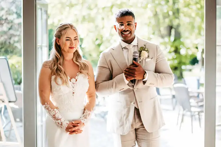 Eine Braut in einem weißen Spitzenkleid und ein Bräutigam in einem beigen Anzug stehen auf ihrer Hochzeit Gut am See. Der Bräutigam hält ein Mikrofon in der Hand und lächelt, während die Braut ihn anschaut. Beide wirken glücklich und werden von natürlichem Licht angestrahlt.