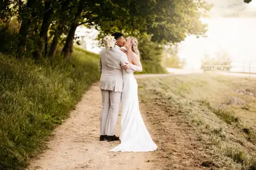 Eine Braut und ein Bräutigam umarmen und küssen sich auf einem von Bäumen gesäumten Feldweg in der Nähe eines Sees bei Hochzeit Gut am See. Die Braut trägt ein langes weißes Kleid, während das Sonnenlicht durch die Bäume fällt und eine romantische Atmosphäre schafft, aufgenommen von Hochzeitsfotograf Görlitz.